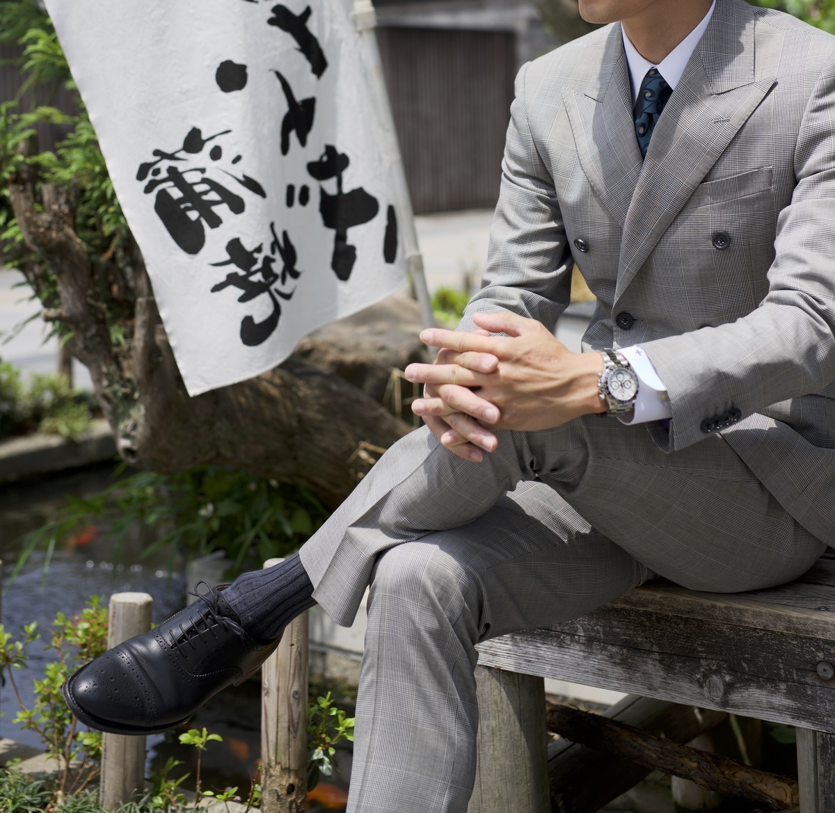 グレーのダブルスーツを着た男性が川沿いのベンチでくつろぐ様子