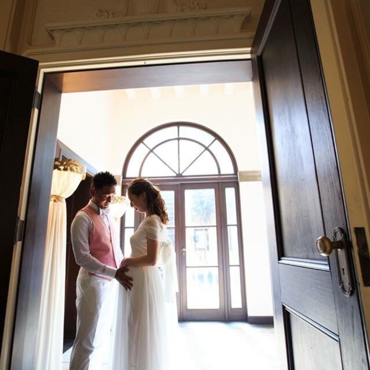 ウェディングドレス姿の新婦と新郎が窓辺の扉の前で向き合う様子